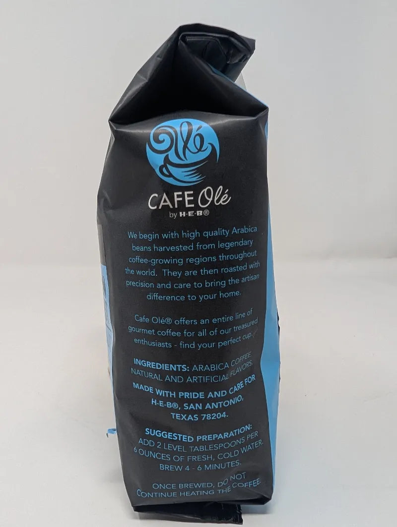Side view of a dark gray and blue 12 ounce bag of Cafe Ole Cinnamon Hazelnut whole bean coffee on a white background featuring an ingredients list.