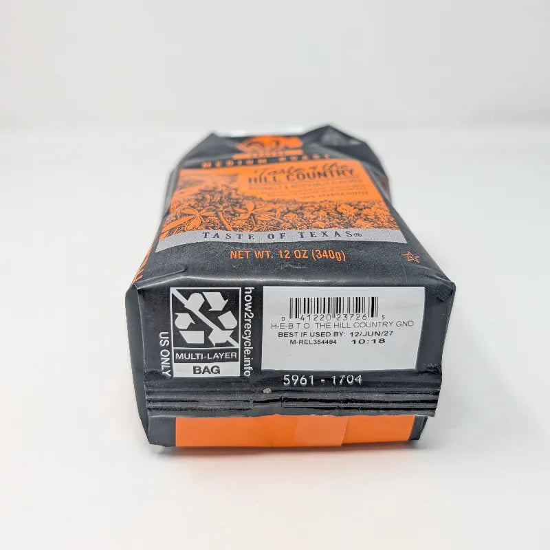 Bottom view of a 12 oz bag of Cafe Ole by H-E-B Ground Medium Roast Taste of the Hill Country coffee, featuring a dark gray and orange design with illustrations of Texas flowers on a white background.