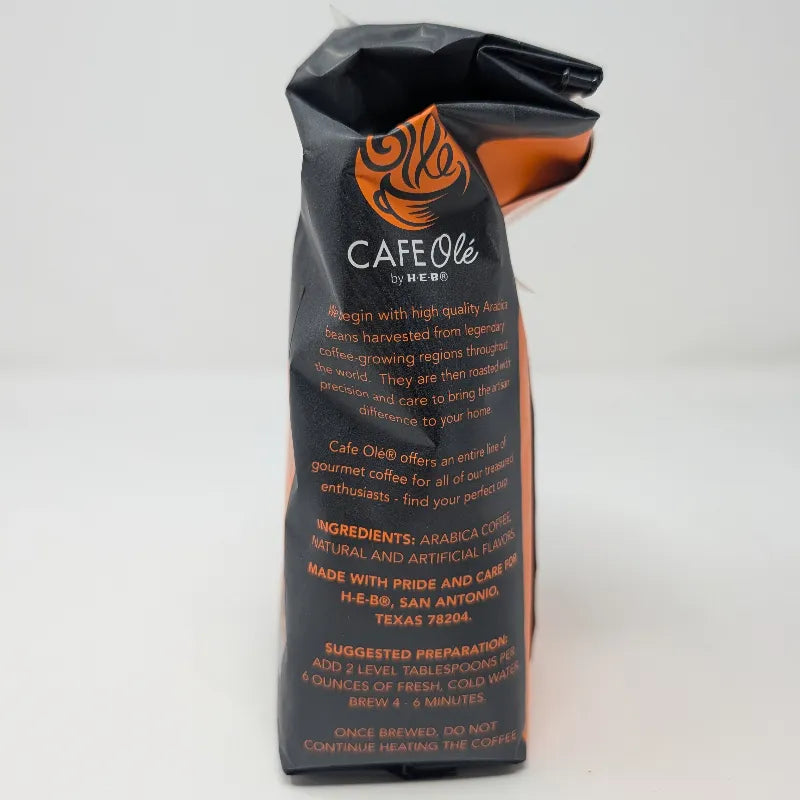 Side view of a 12 oz bag of Cafe Ole by H-E-B Ground Medium Roast Taste of the Hill Country coffee, featuring a dark gray and orange design with ingredients list on a white background.