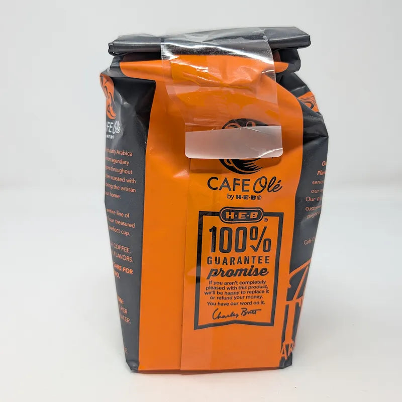 Rear view of a 12 oz bag of Cafe Ole by H-E-B Ground Medium Roast Taste of the Hill Country coffee, featuring a dark gray and orange design on a white background.