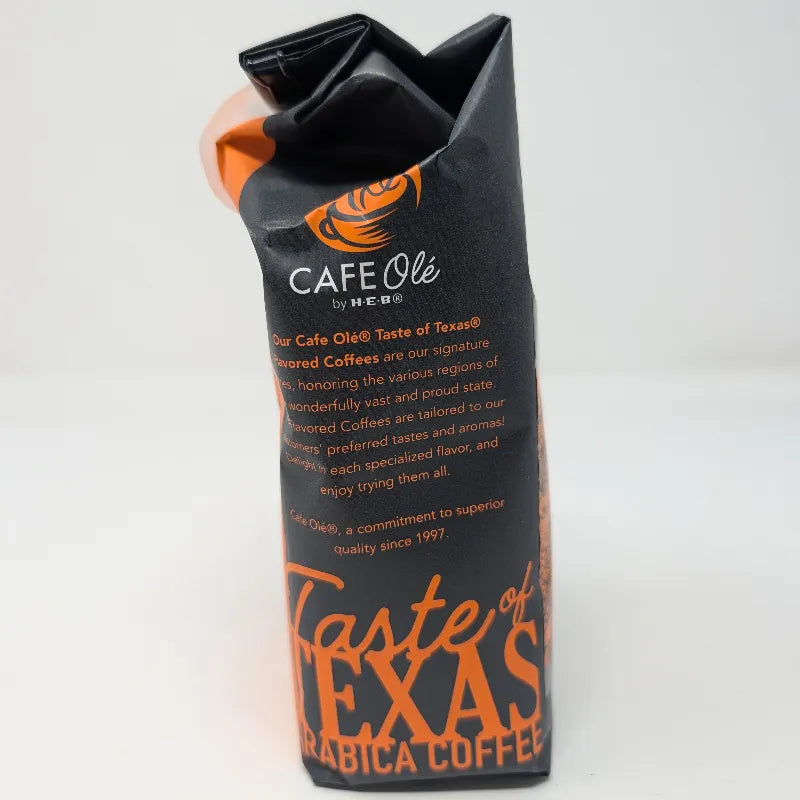Side view of a 12 oz bag of Cafe Ole by H-E-B Ground Medium Roast Taste of the Hill Country coffee, featuring a dark gray and orange design on a white background.