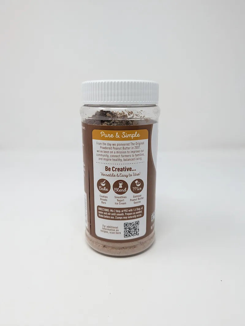 Side view 2 of jar of PB2 Cocoa Peanut Powder on a plain white background. The product is in a clear plastic jar showing the brown powder inside, with a white ribbed lid and a brown and white label featuring the PB2 logo and cocoa bean illustrations.
