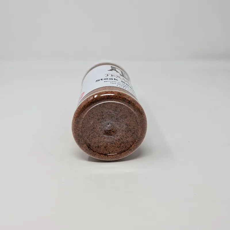 Bottom view of a clear plastic bottle of Texas A&M University Steak Seasoning with a white cap and white label, containing brown seasoning powder, on a white background.