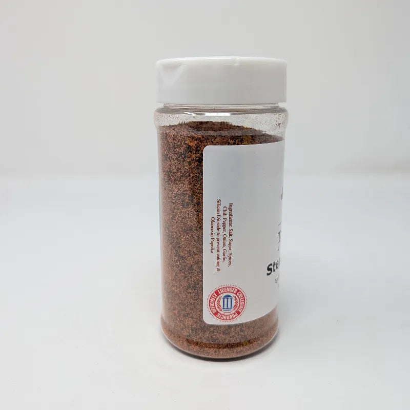 Side view of a clear plastic bottle of Texas A&M University Steak Seasoning with a white cap and white label including an ingredients list, containing brown seasoning powder, on a white background.